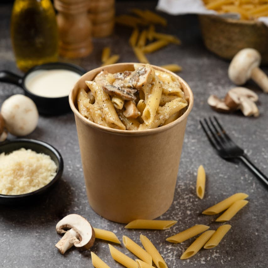 Pasta dish in a paper cup with ingredients on a dark surface