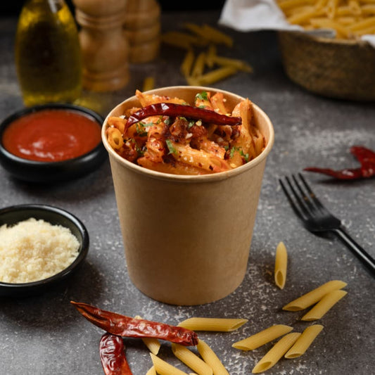 Paper cup with pasta and sauce on a dark surface with ingredients around