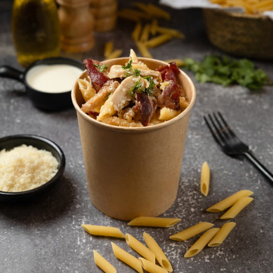 Pasta dish in a paper cup on a table with ingredients and utensils.