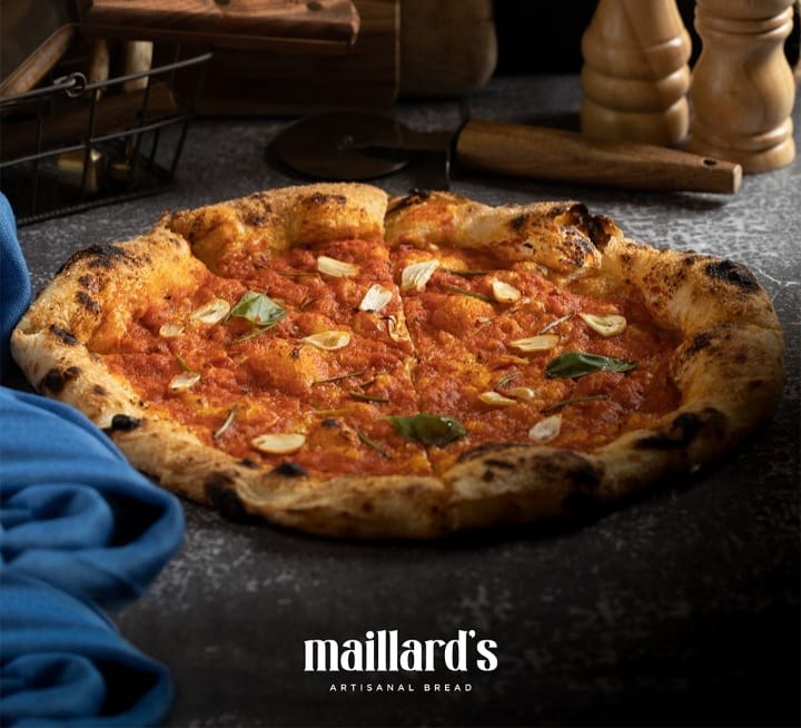 Pizza with tomato sauce and green leaves on a dark surface, featuring the Maillard's logo.